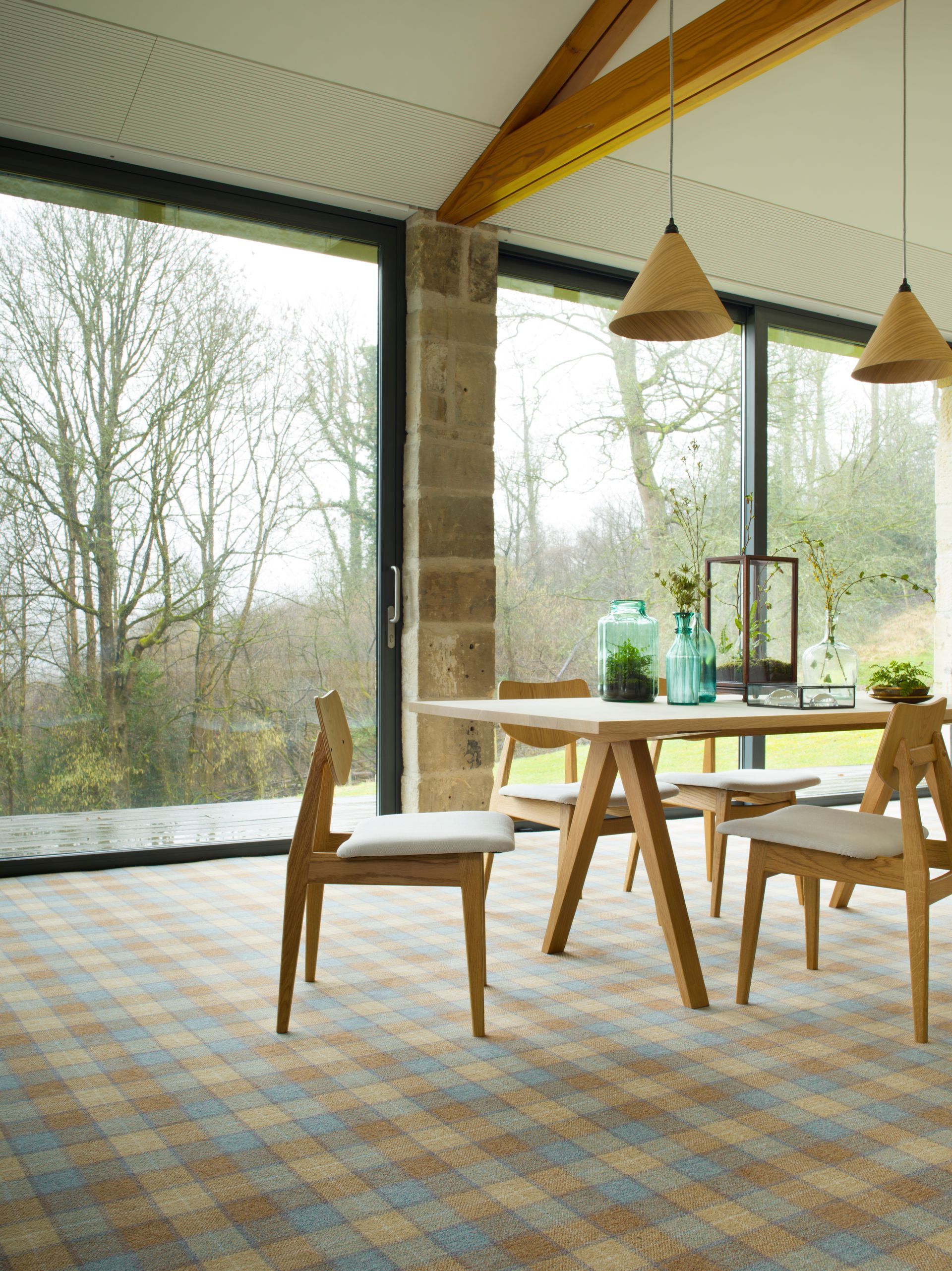 Dining area with a wooden table, chairs, and plaid carpet, featuring floor-to-ceiling windows overlooking a forest.