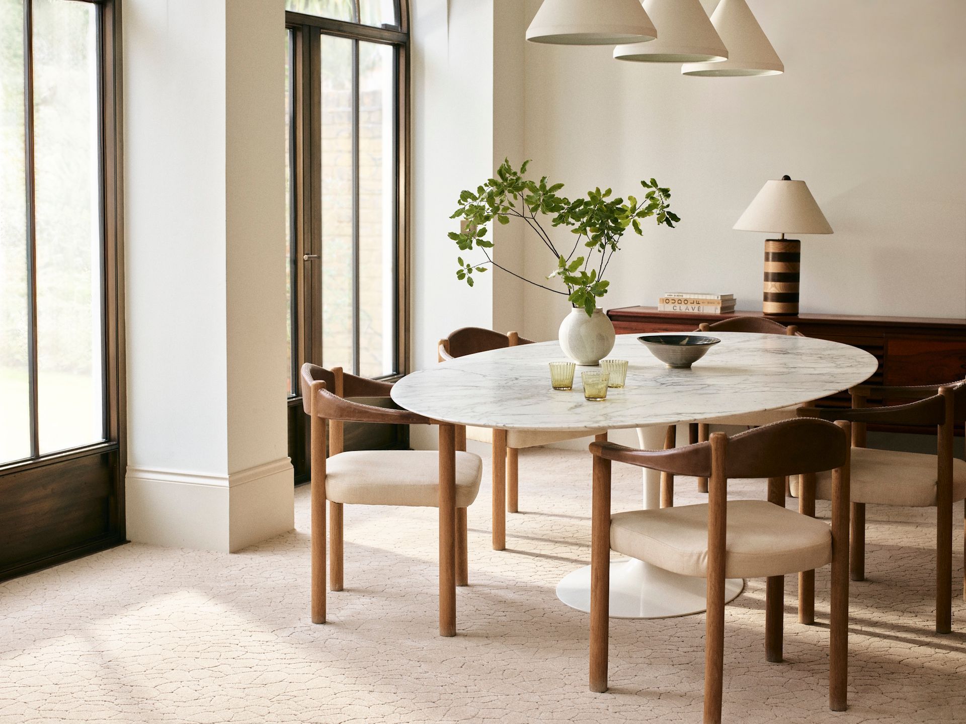A dining room featuring an oval marble table with four wood and fabric chairs, lit by three hanging pendant lamps.