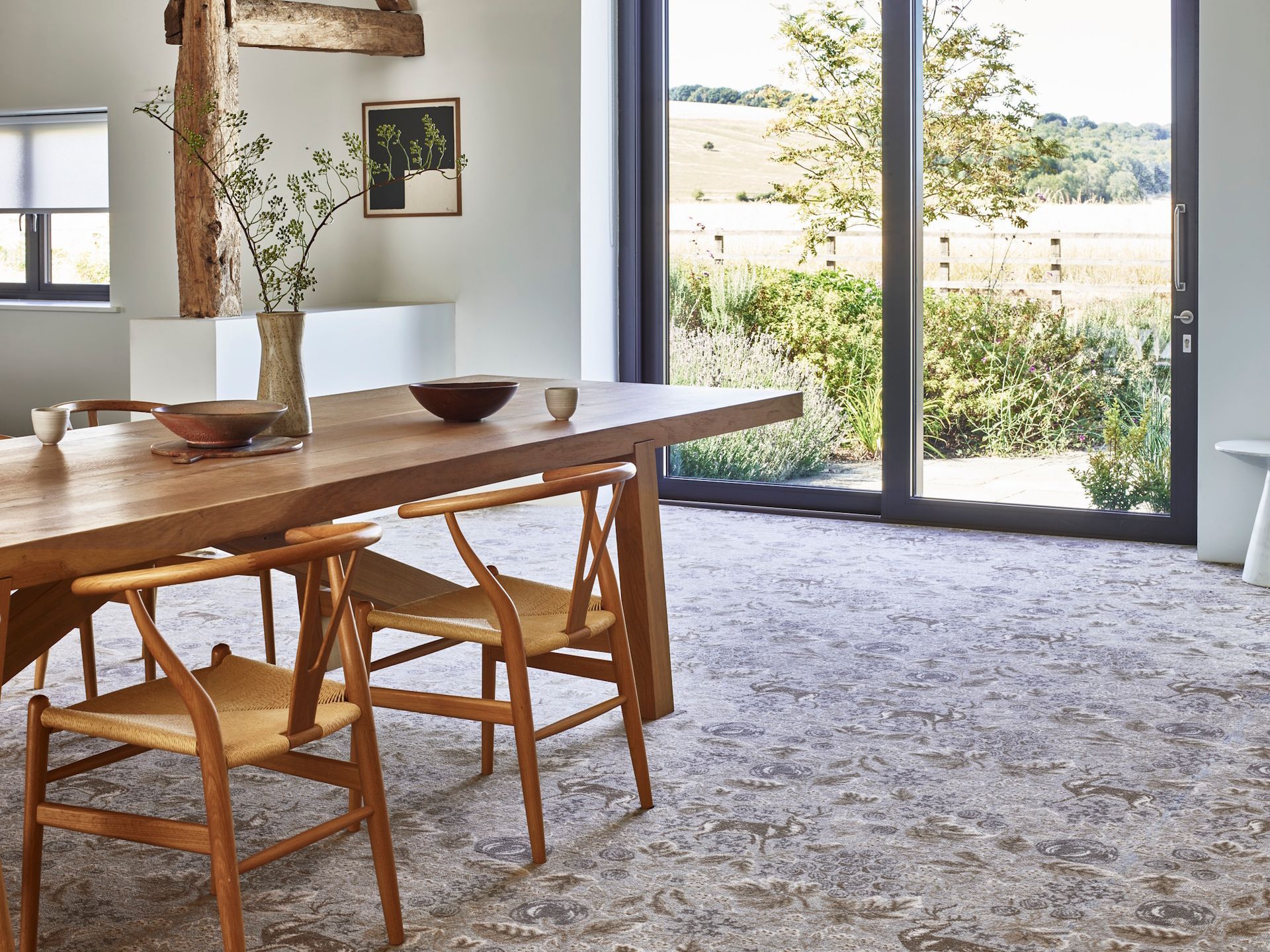 A light wood dining table with woven-seat chairs sits in a bright, modern room next to a large glass door overlooking fields.