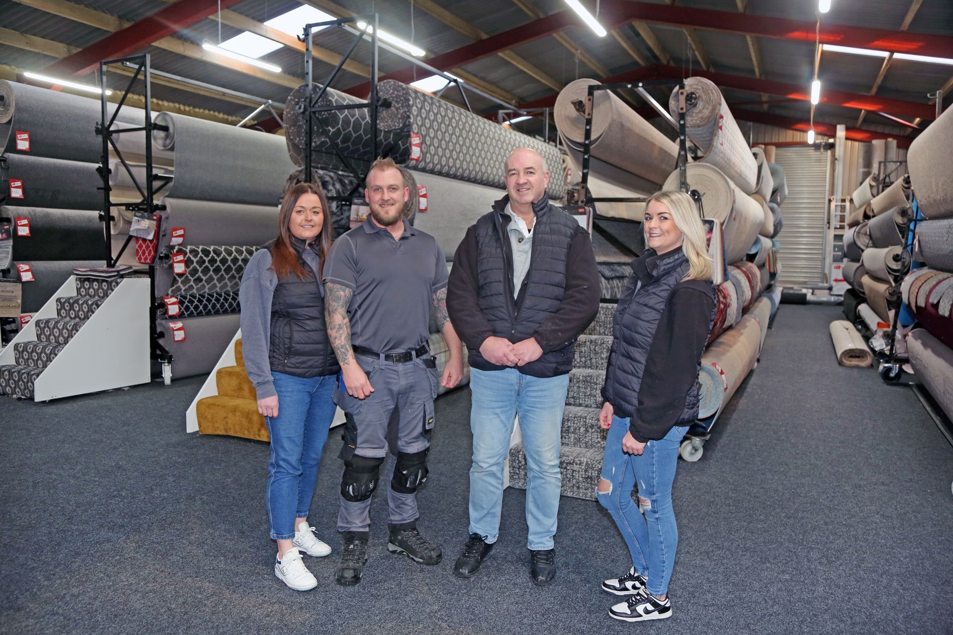Four people stand in a warehouse aisle filled with stacked rolls of carpet, posing for a group photograph.
