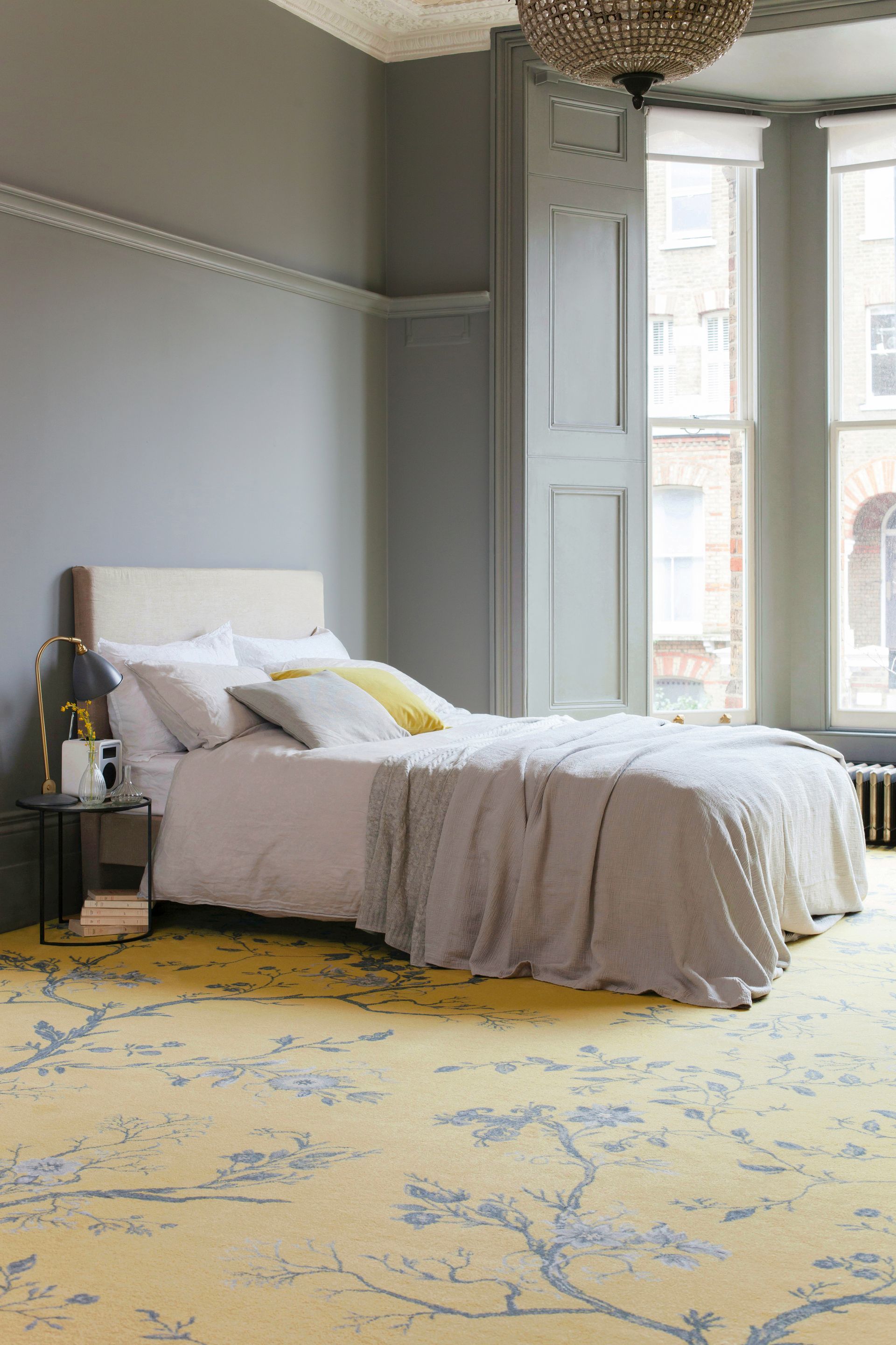 A bedroom with gray walls and tall windows, featuring a bed with neutral linens and a yellow floral-patterned rug.