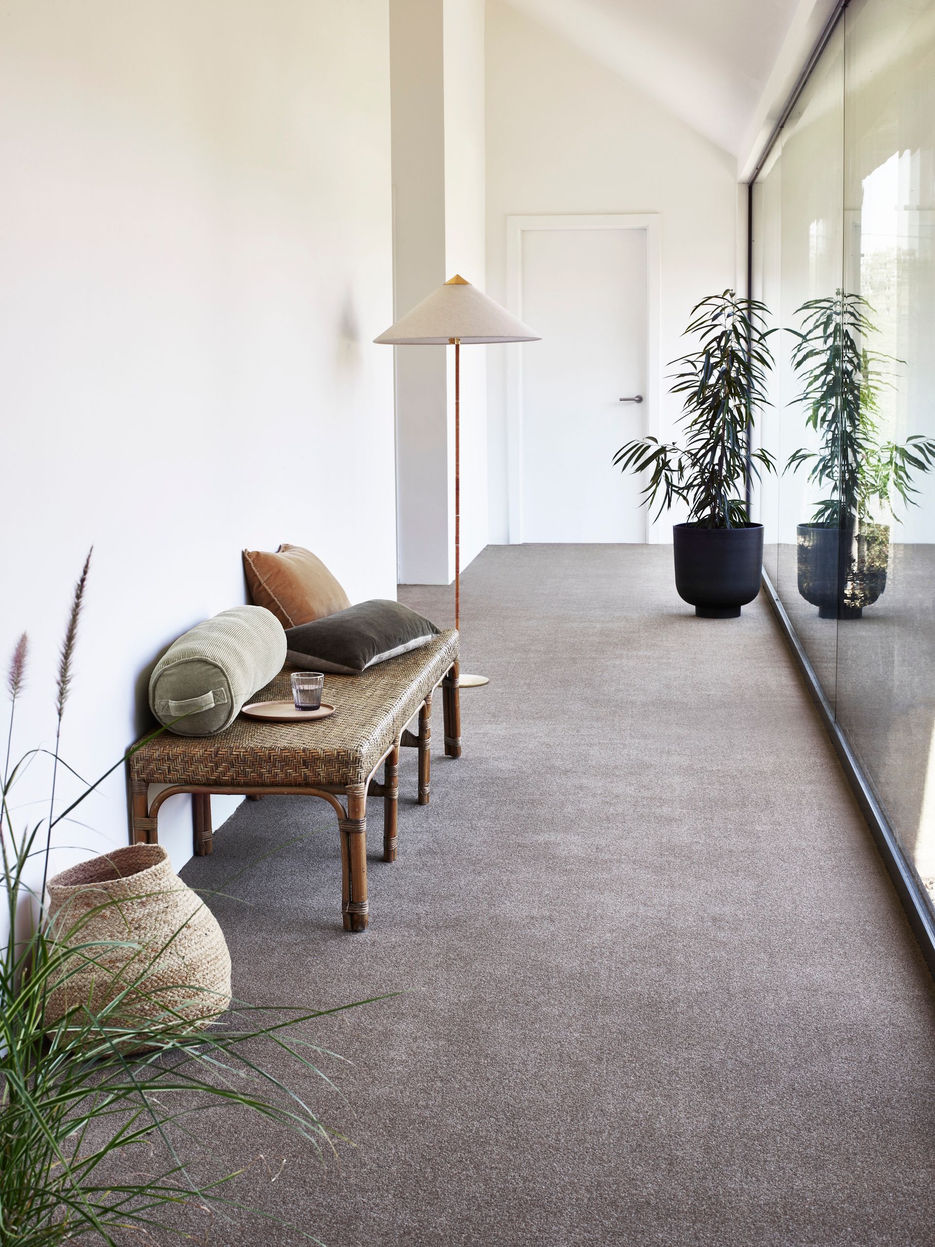A hallway with a woven bench, textured cushions, a floor lamp, and two potted plants against a window wall.