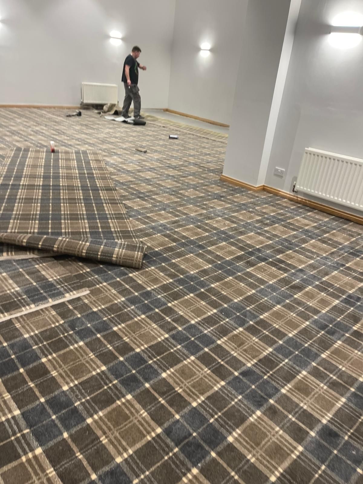Room with plaid carpet being installed; person standing, working; neutral colors, bright lighting.