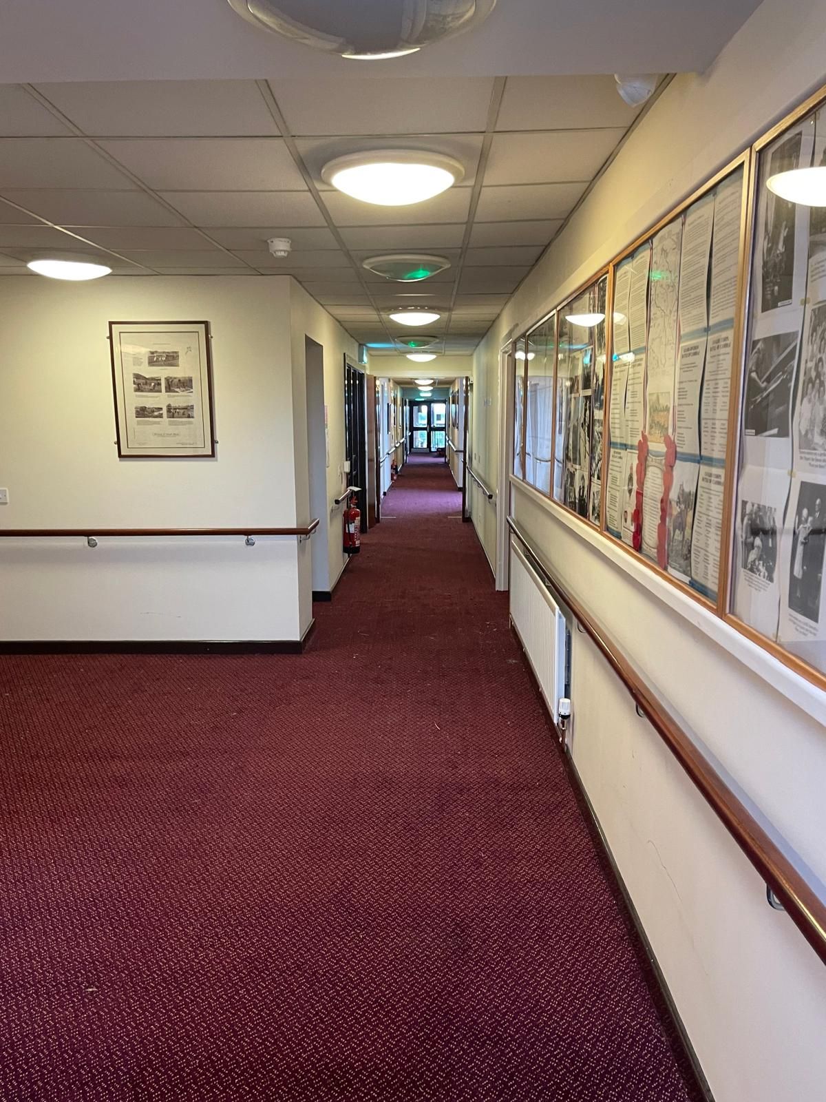 Long hallway with red patterned carpet, handrails, and framed displays on one side, lit by ceiling lights.