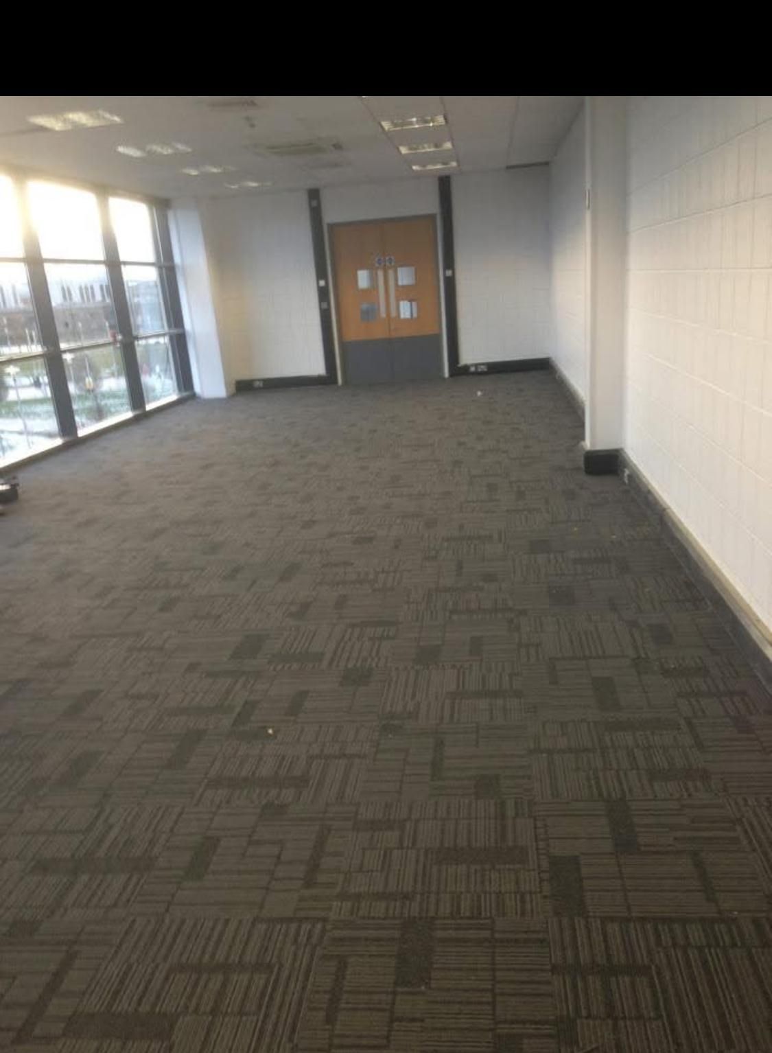 Empty hallway with patterned dark carpet, large windows on left, door at end, white walls.