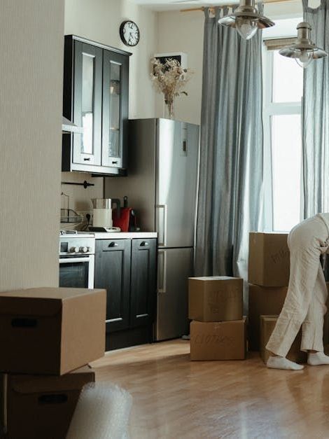 A woman is kneeling down in front of a stack of cardboard boxes in a kitchen.