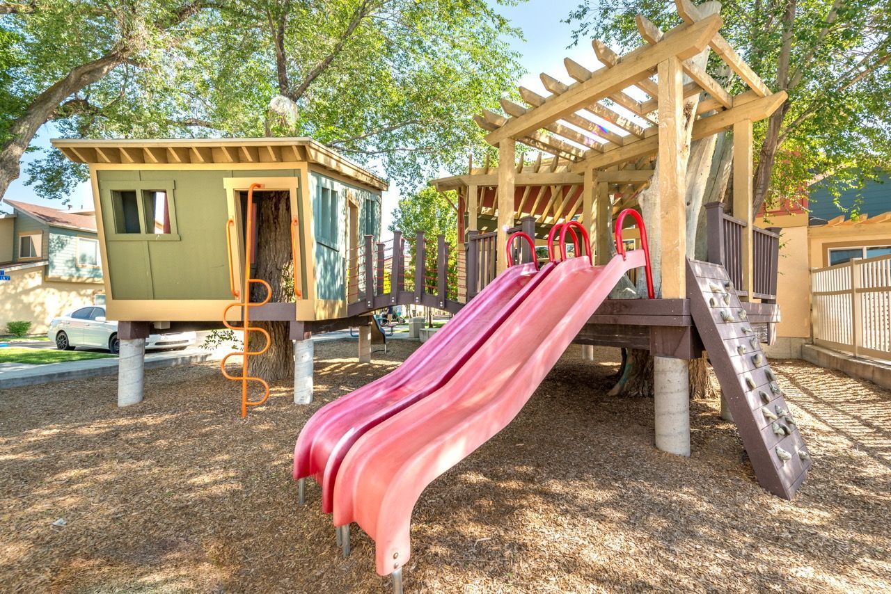 Playground at an apartment community with a wooden play structure and pink slides under trees.