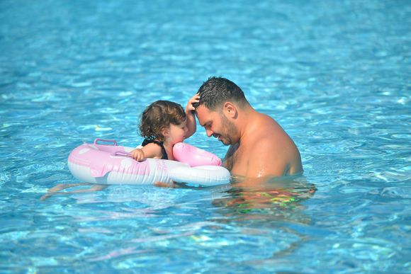 An adult and a child in an inflatable ring swim in a bright blue pool, with the child touching the adult&rsquo;s head.