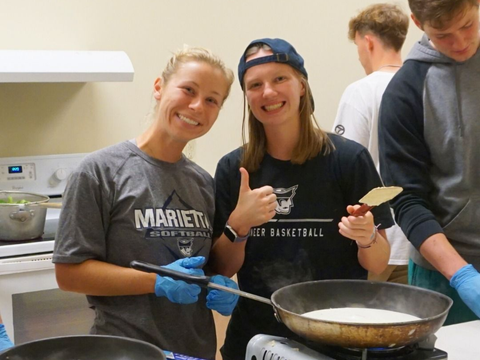 Two people in a kitchen, one wearing a Marietta Softball shirt and the other giving a thumbs-up while cooking.