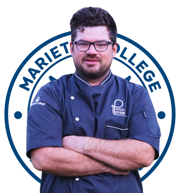 Man in chef's uniform with crossed arms, wearing glasses. The emblem of a culinary institute is in the background.