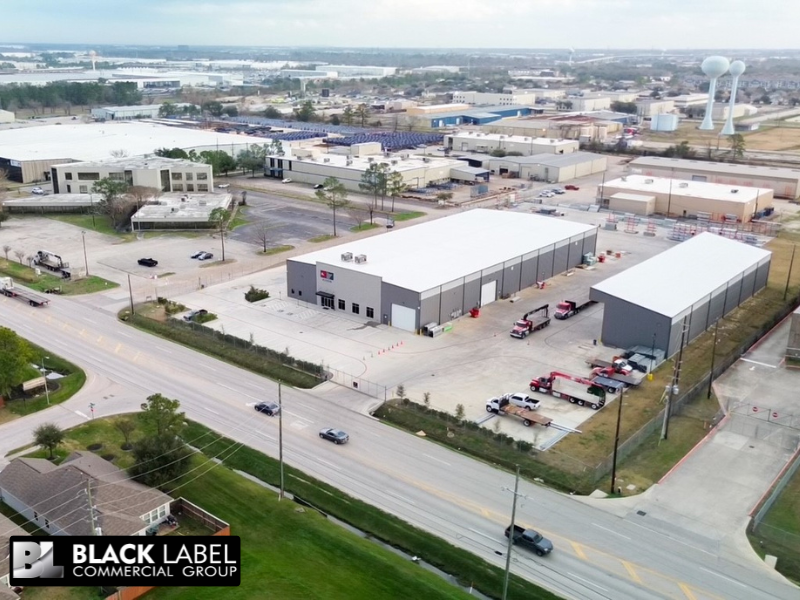 Aerial view of two gray industrial buildings with vehicles parked outside. Suburban setting.