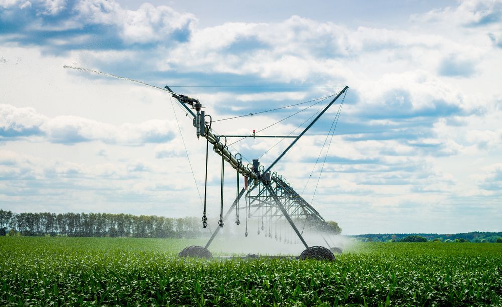 Center-pivot irrigation system spraying water over a green field of crops under a cloudy sky. — Bennett's Servicing and Plumbing In South Nowra, NSW