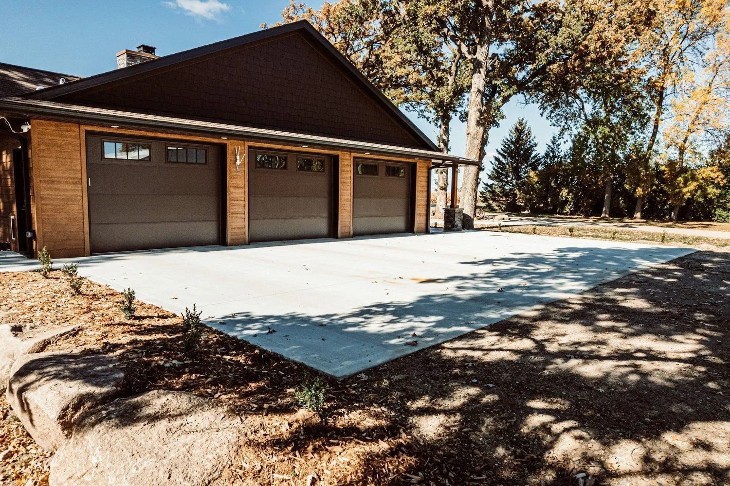 Three-car garage with gray doors, tan brick siding, and a concrete driveway, set against a backdrop of trees.