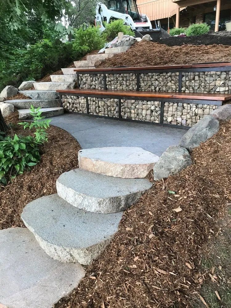 Stone steps and tiered seating built into a hillside, using stone-filled wire baskets and wooden benches.