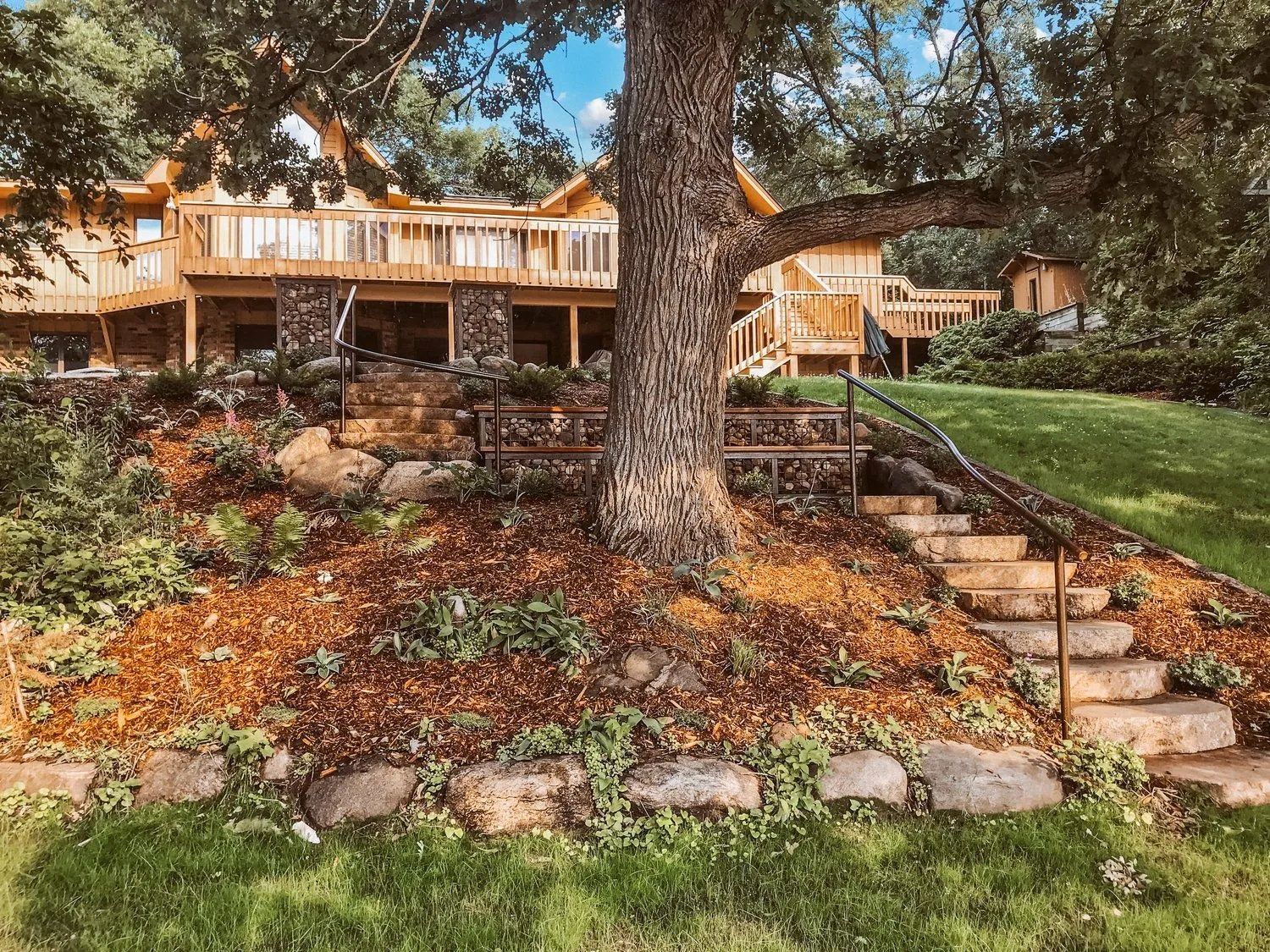 Treehouse with wooden decks, stone steps, and a grassy hill on a sunny day.