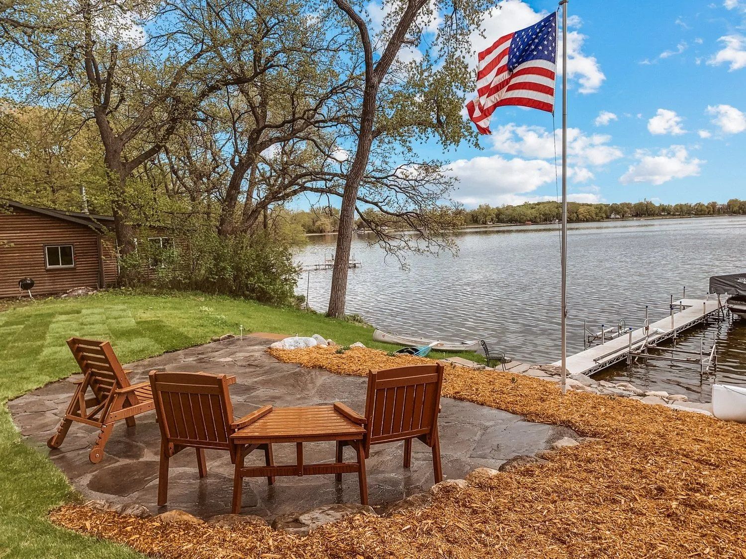 Patio with wooden chairs, table, and American flag overlooking a lake.
