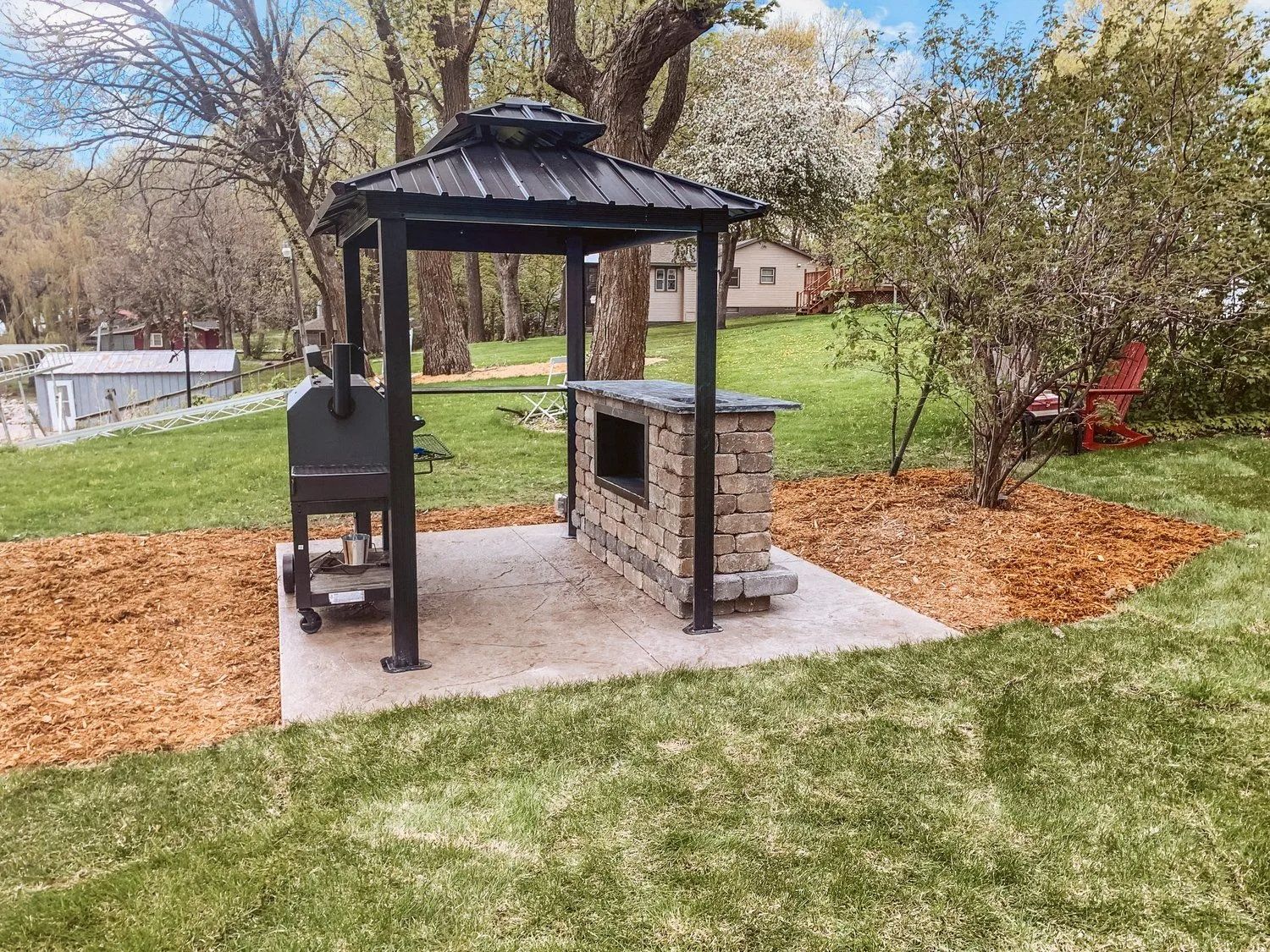 Outdoor kitchen with black metal gazebo, stone-faced bar, grill on concrete pad, and wood chips in yard.