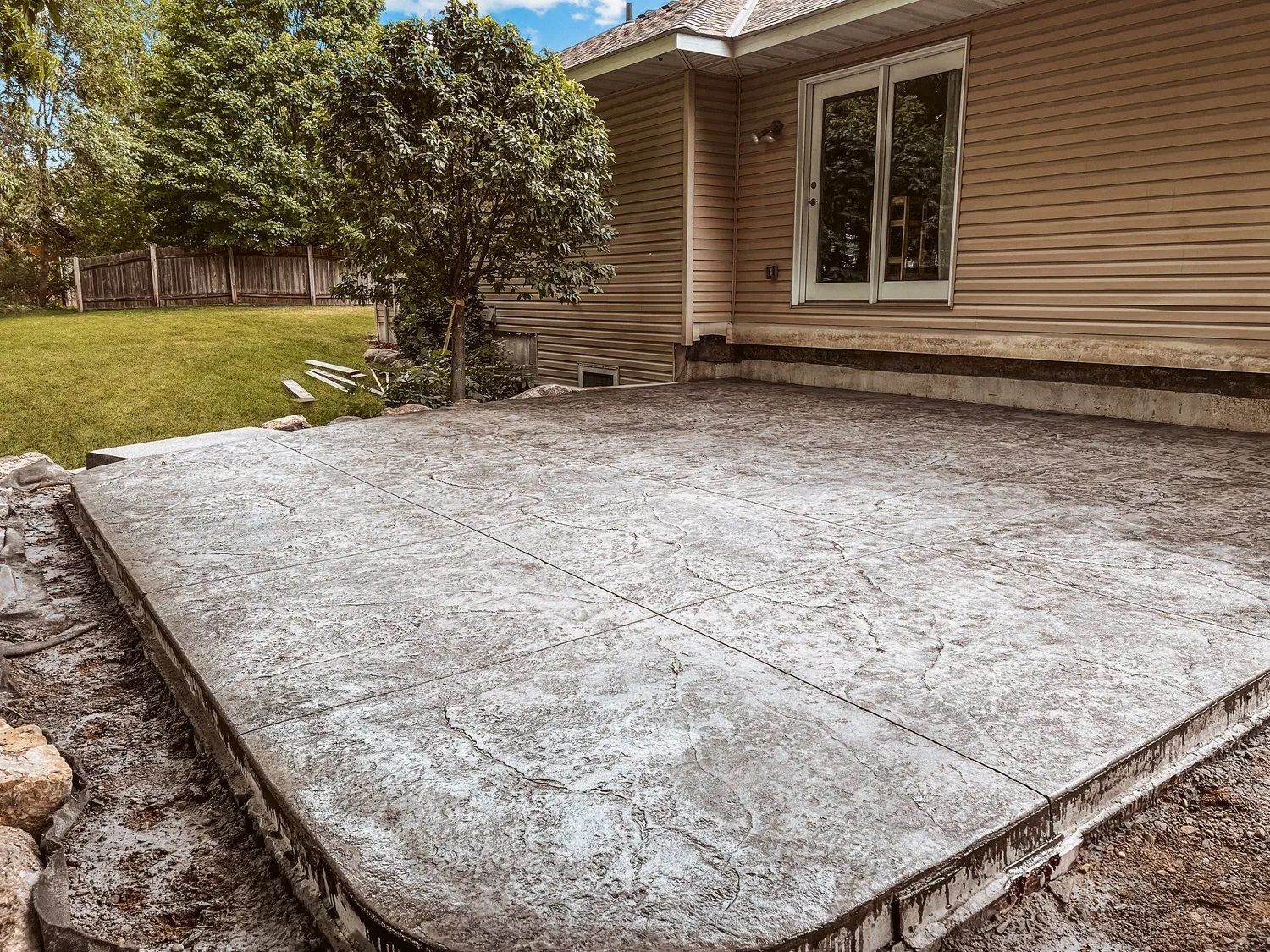Concrete patio with a house in the background. Grass and a tree are also visible.