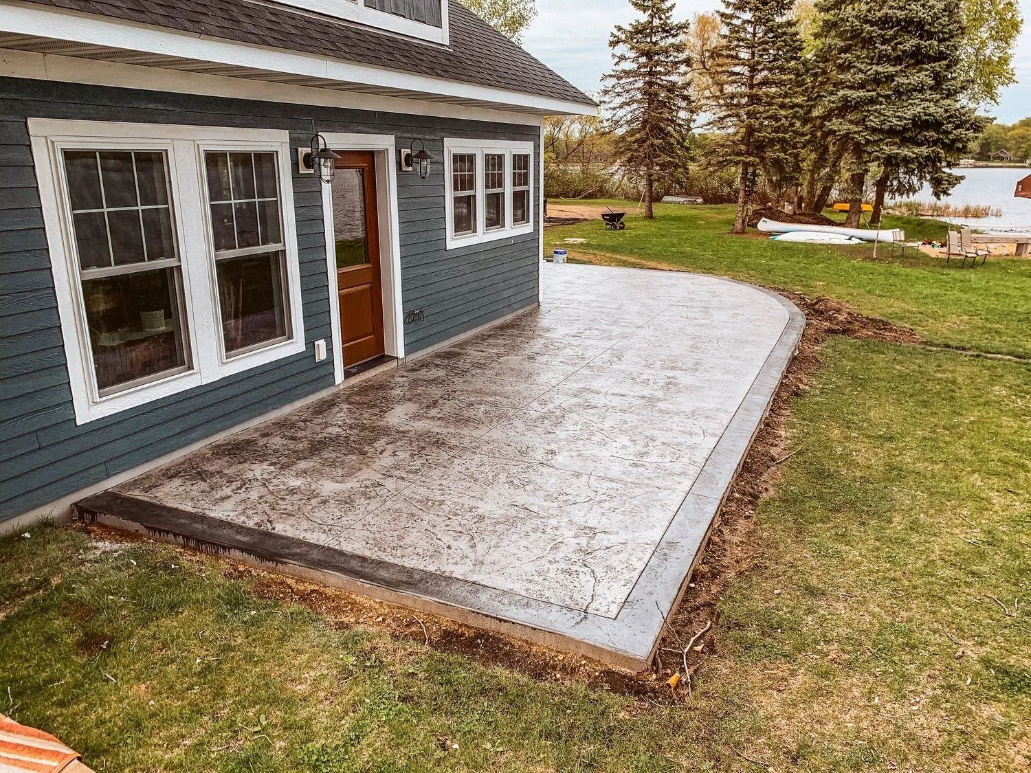 Concrete patio with blue house and brown door, on green grass near a body of water.