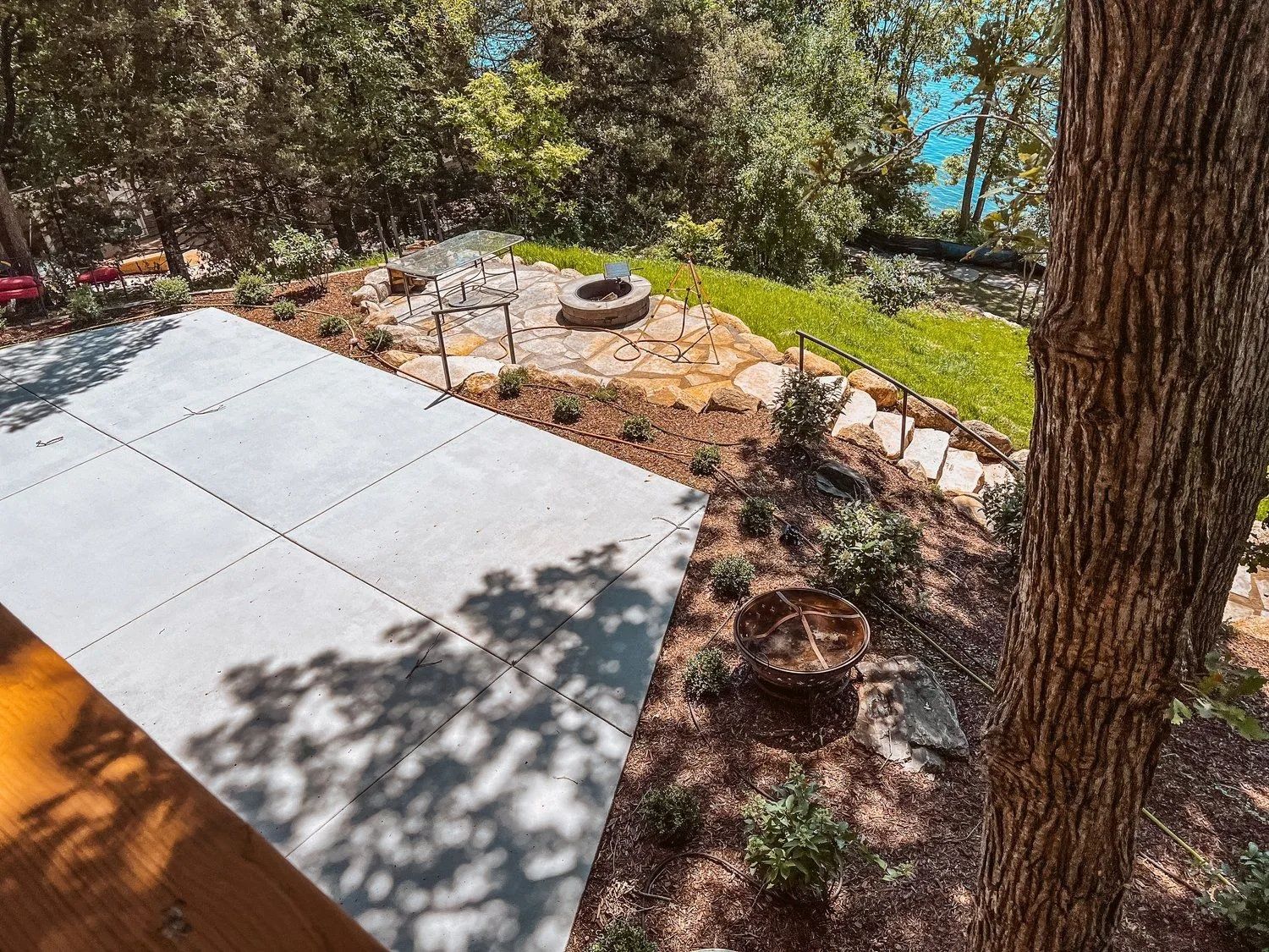 Patio area with fire pit, stone path, and tree, overlooking a lake and trees.