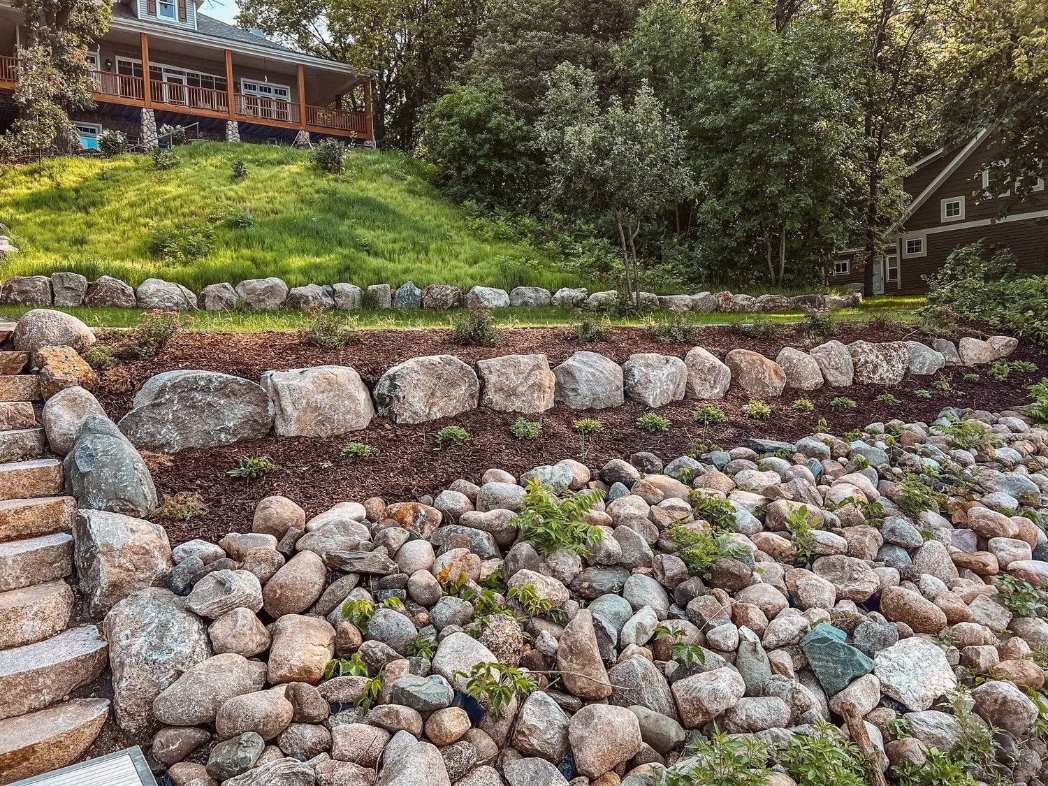 Stone retaining wall with a house on a grassy hill and a house in the background.