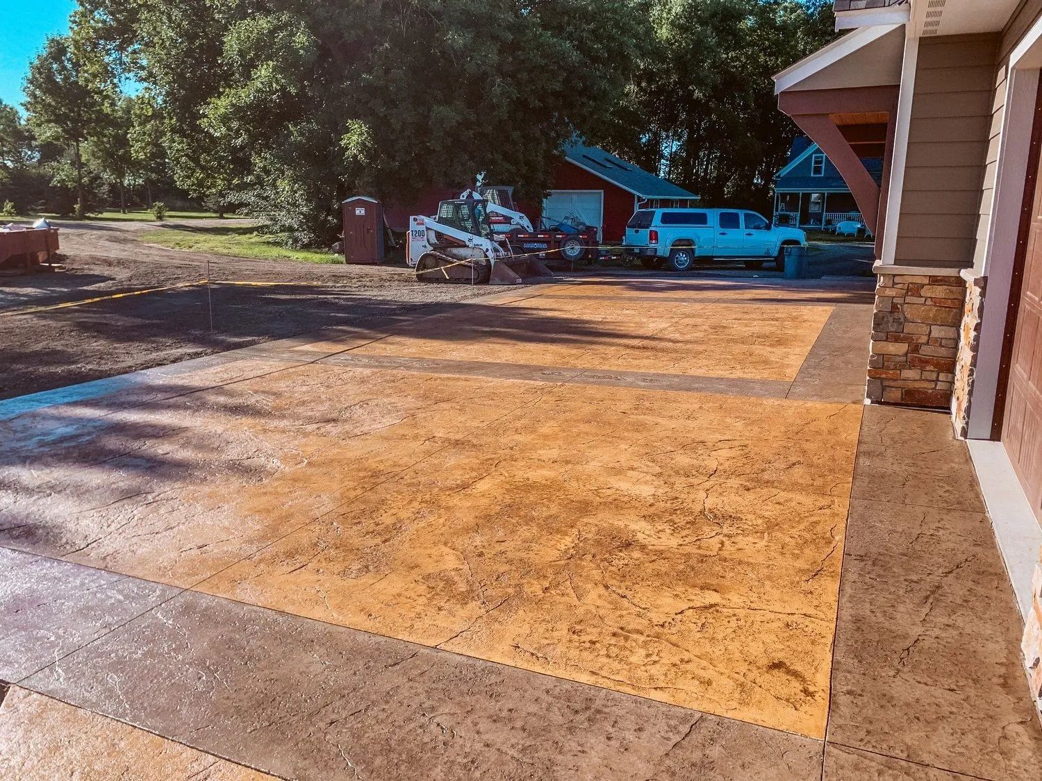 Concrete driveway with a brown border and tan center, construction vehicles and buildings in the background.