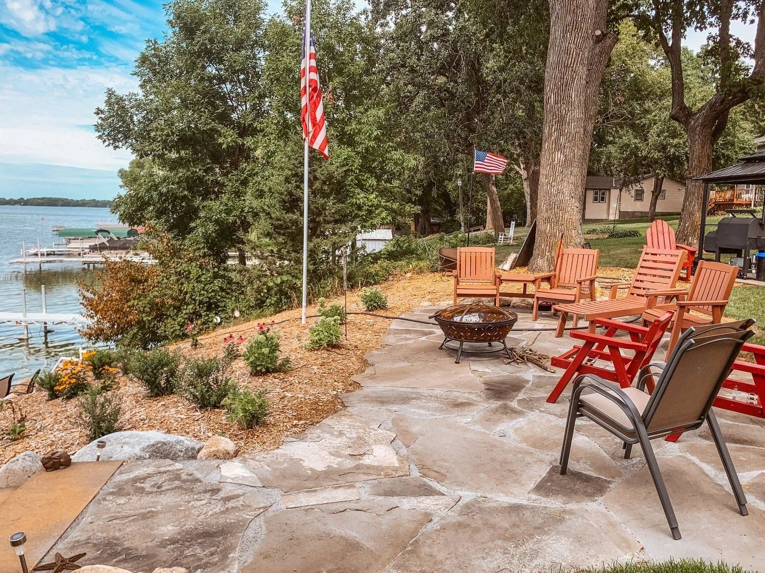 Lakeside patio with red Adirondack chairs, fire pit, American flag, and water view.