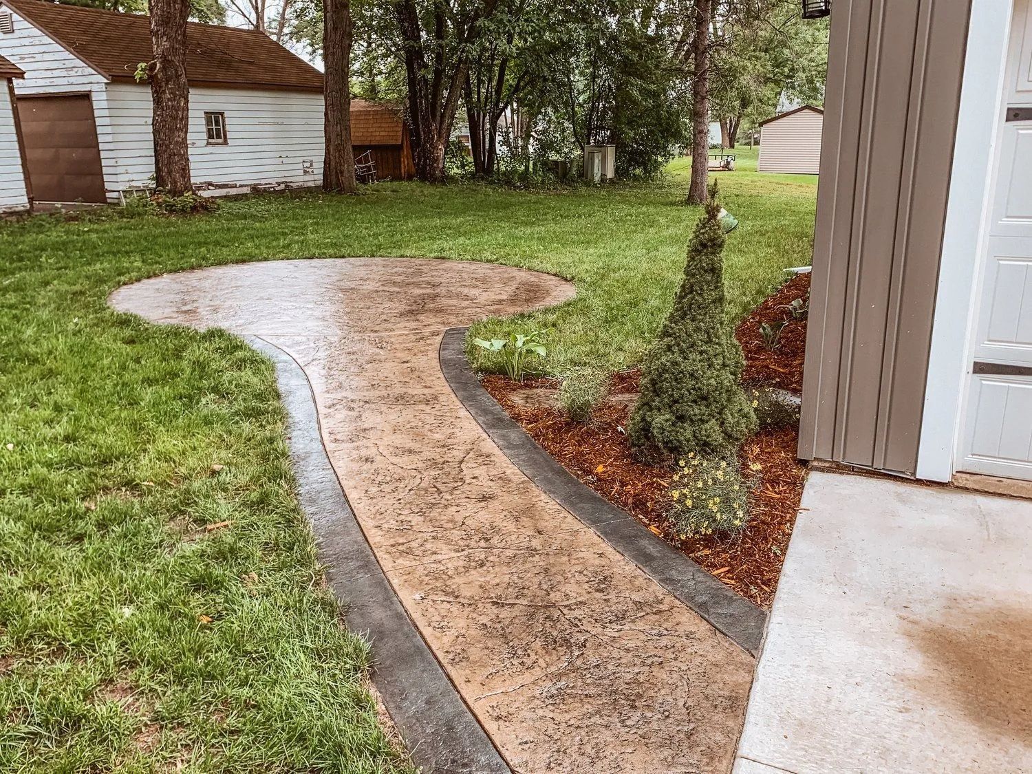Stamped concrete pathway with a circular patio area, bordered by dark concrete and a bed of mulch, leading from a building.