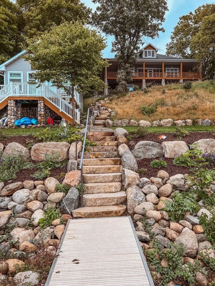 Stone steps lead up to two houses on a hillside, lined with rocks and greenery, with a wooden dock in the foreground.
