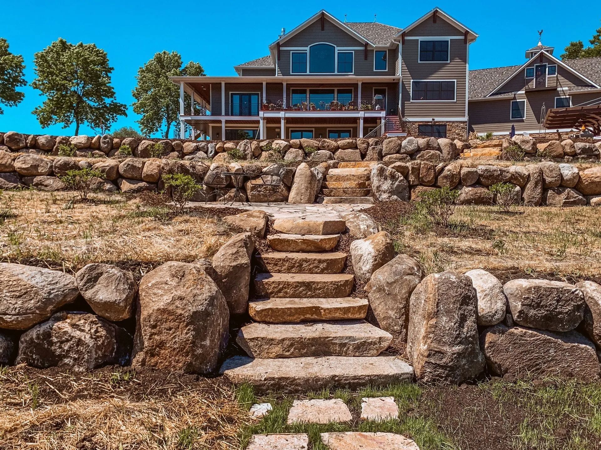 Stone steps lead up to a large house with a porch and multiple windows, under a blue sky.