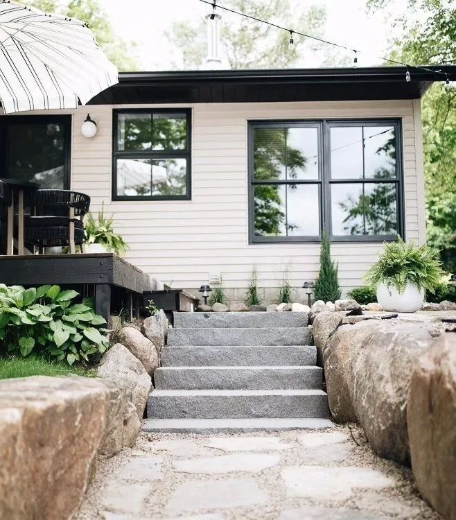 Stone steps lead to a light-colored building with black-framed windows and a deck, surrounded by greenery.