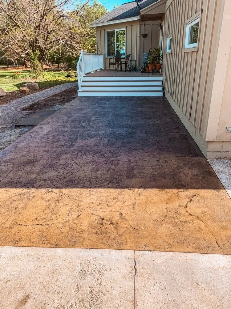 Brown stamped concrete patio next to a light-colored house with a deck. Adjacent to a lighter-colored concrete sidewalk.