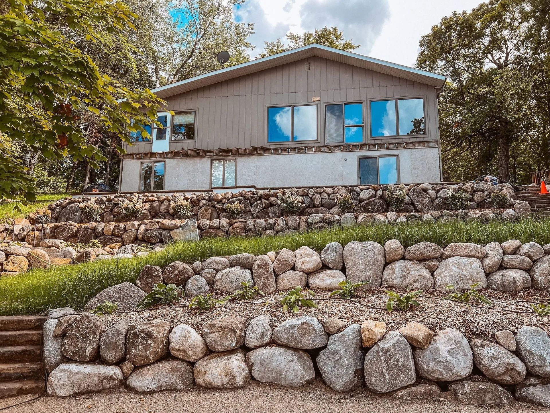 Two-story house with light grey siding, large windows, and rock retaining walls. Green grass and trees surround it.