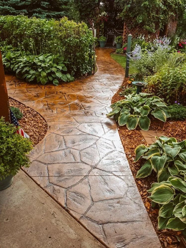 Concrete path through a garden with plants, bordered by mulch.