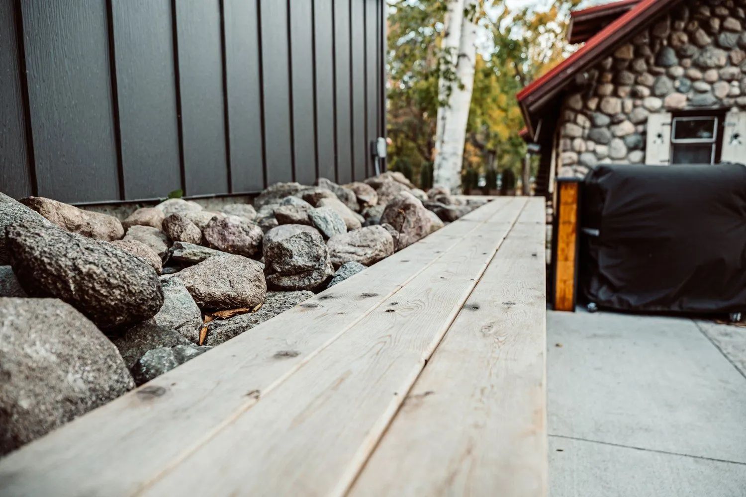 Wooden walkway next to a rock garden and building with dark siding and stone exterior.