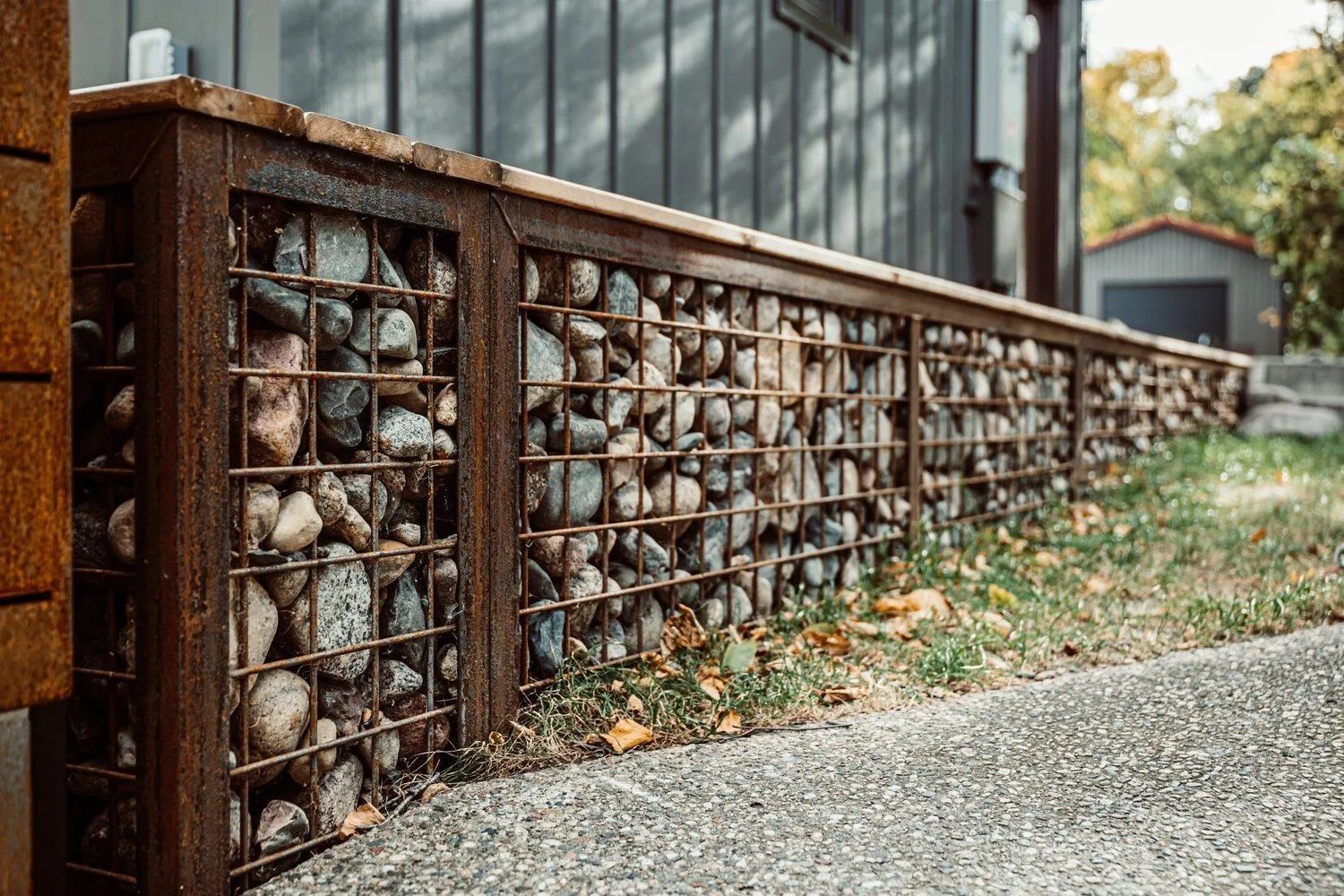 Gabion wall filled with rounded stones along a paved walkway. Weathered brown metal frame, wood top.