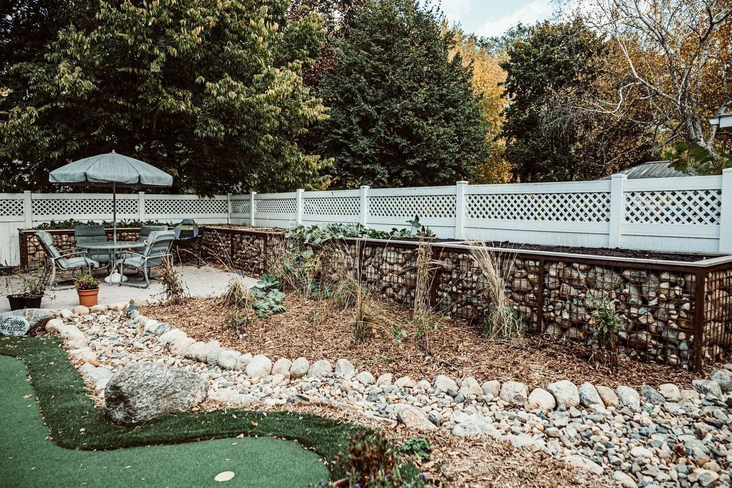 Backyard with stone garden beds, white fence, patio furniture, and trees.