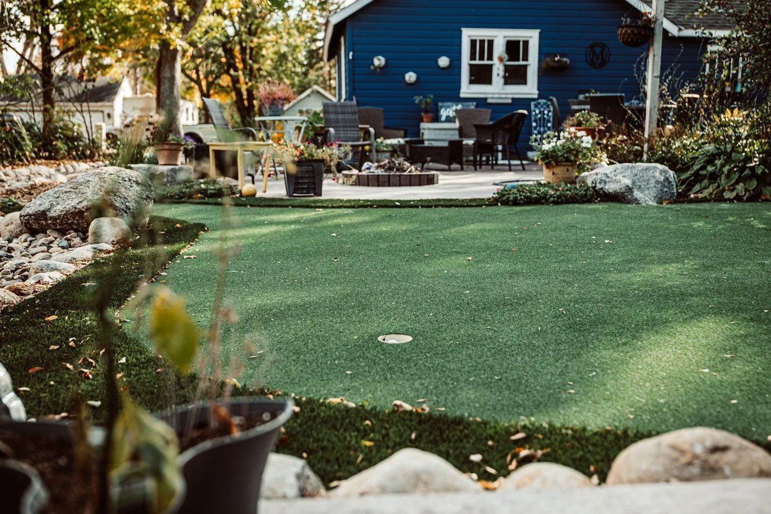 Backyard with blue house, putting green, patio furniture, and potted plants.