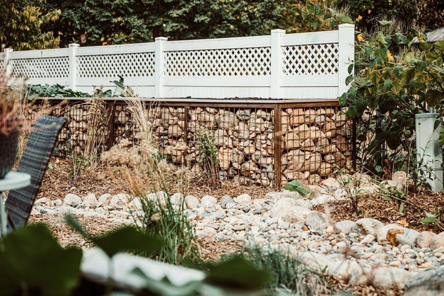 Gabion wall filled with stones, topped with a white lattice fence. A garden with plants and small rocks in the foreground.