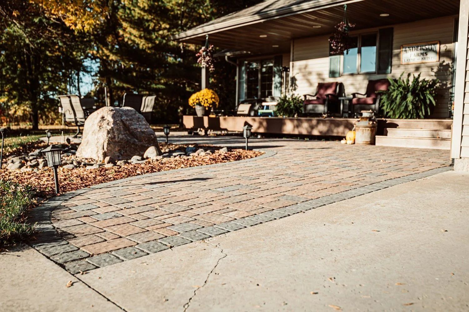 Brick patio in front of a house, featuring a large rock and landscaping.