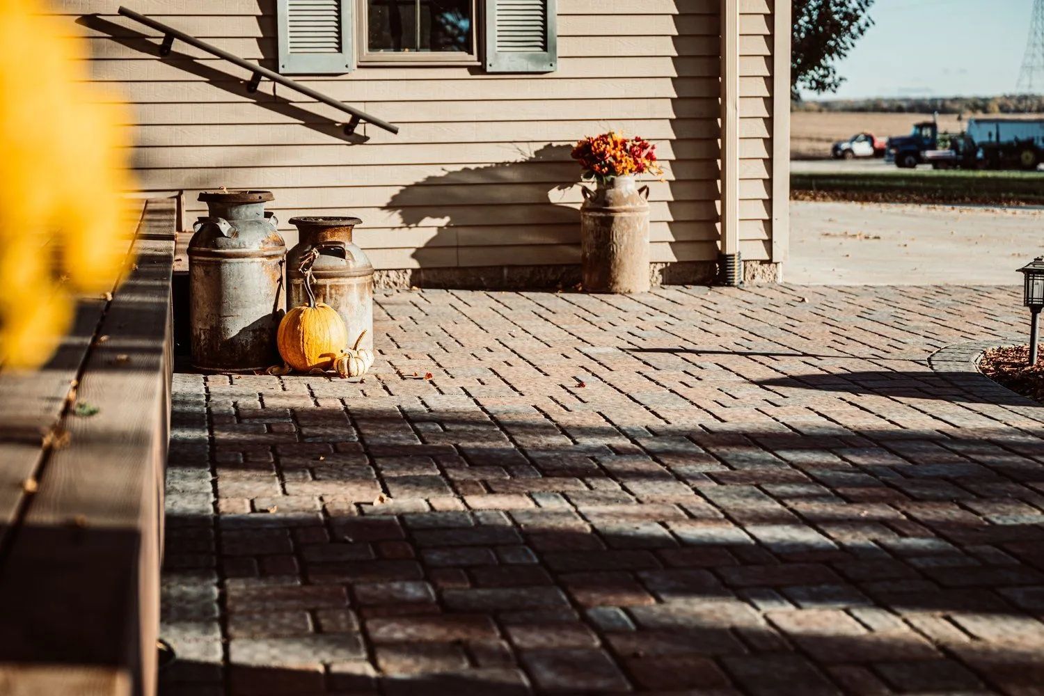 Paver patio with three metal milk cans, a pumpkin, and flowers against a weathered building exterior.