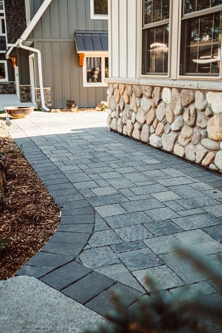 Stone paver pathway leading to a house with stone and wood siding, bordered by mulch and greenery.