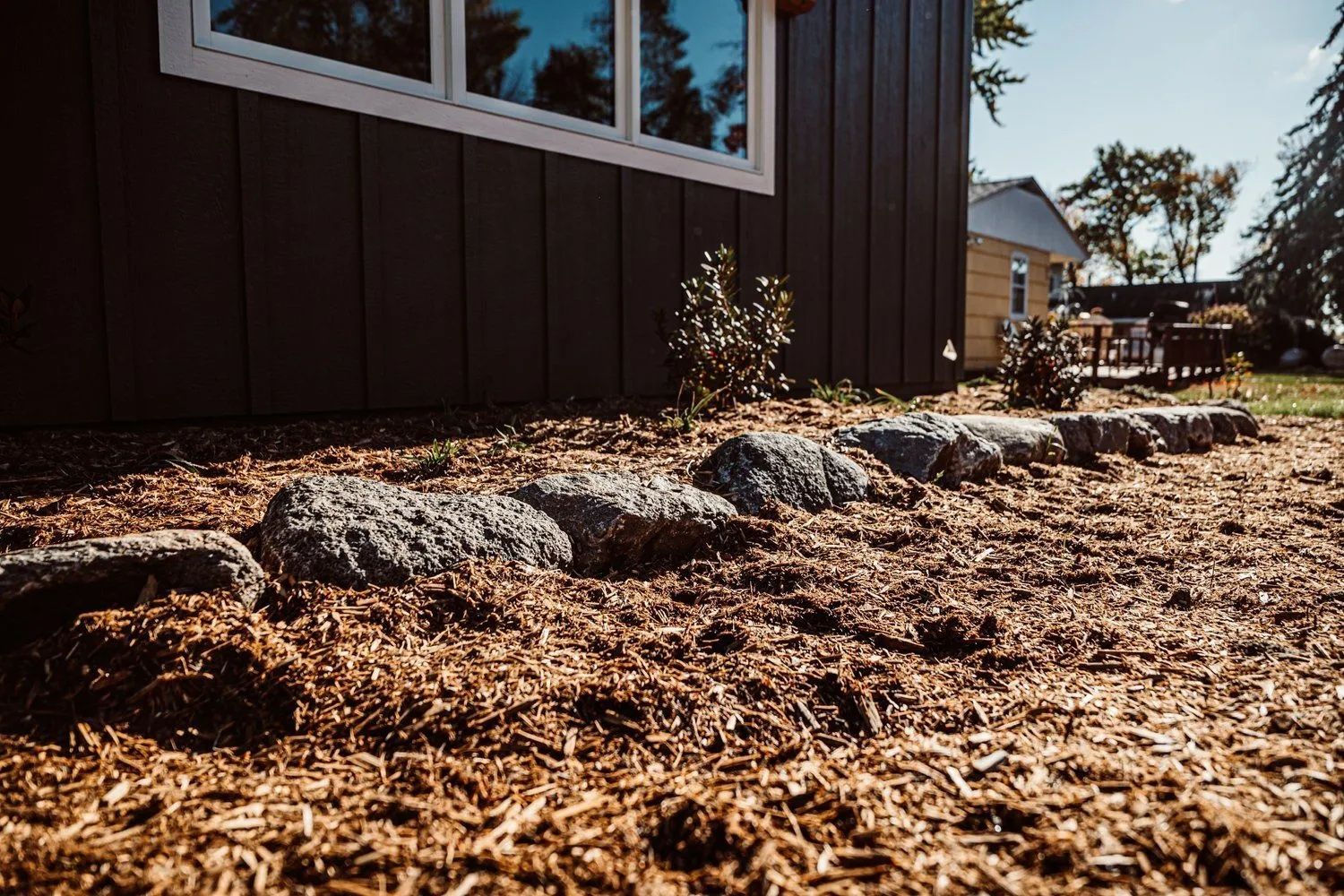 Dark siding of a house with a row of rocks in mulch along the foundation; another house in the background.