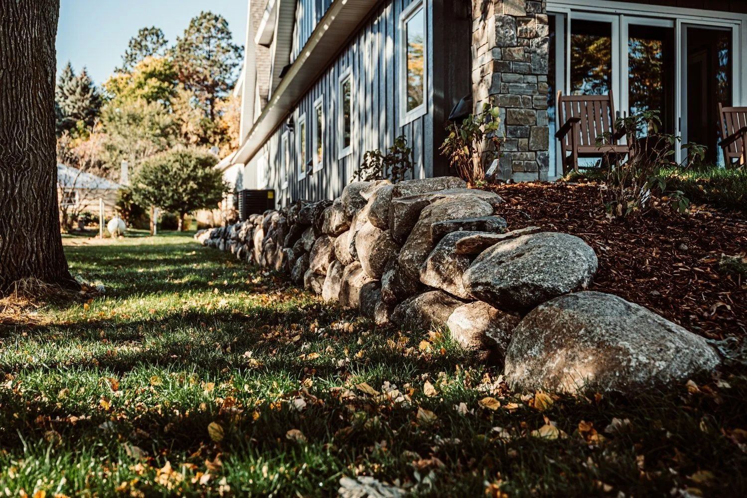 Stone retaining wall next to a house with grass lawn, autumn foliage and trees.