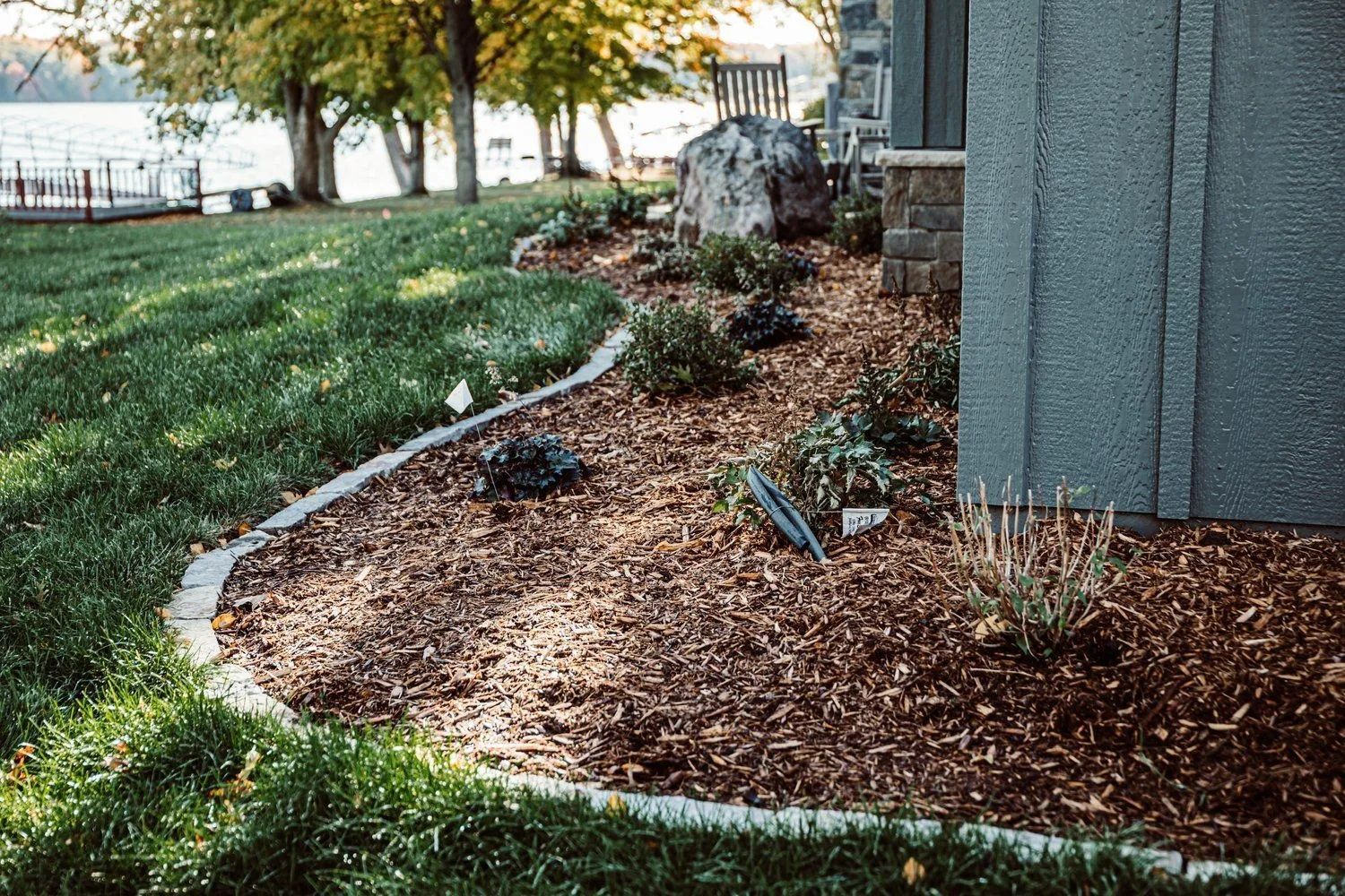 A landscaped garden bed with wood chips and plants, bordered by stone edging, next to a building and grass.