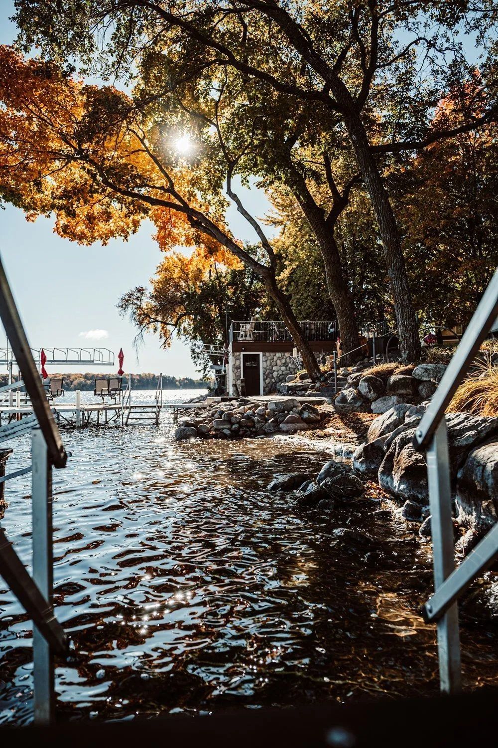 Waterfront scene with stairs leading into the water, with a small stone building and colorful trees on the shore.
