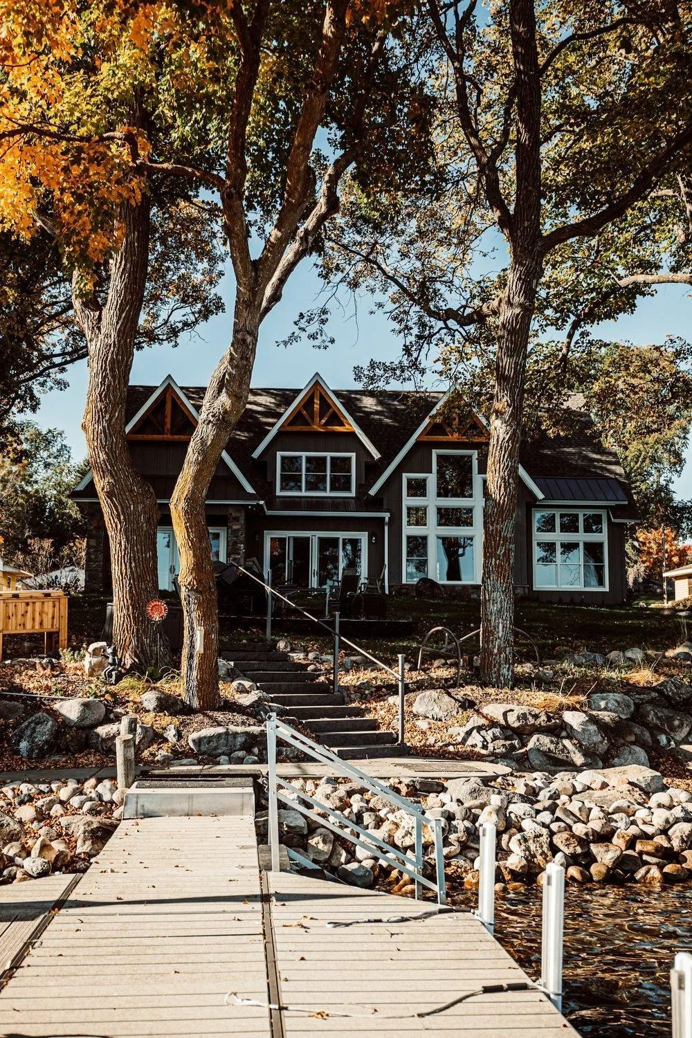 Wooden dock leads to a house by the water, surrounded by trees with fall colors.