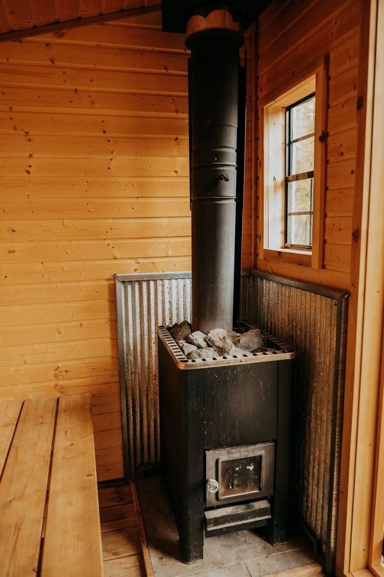 Interior of a sauna with a black stove and chimney, wooden walls, a bench, and a small window.