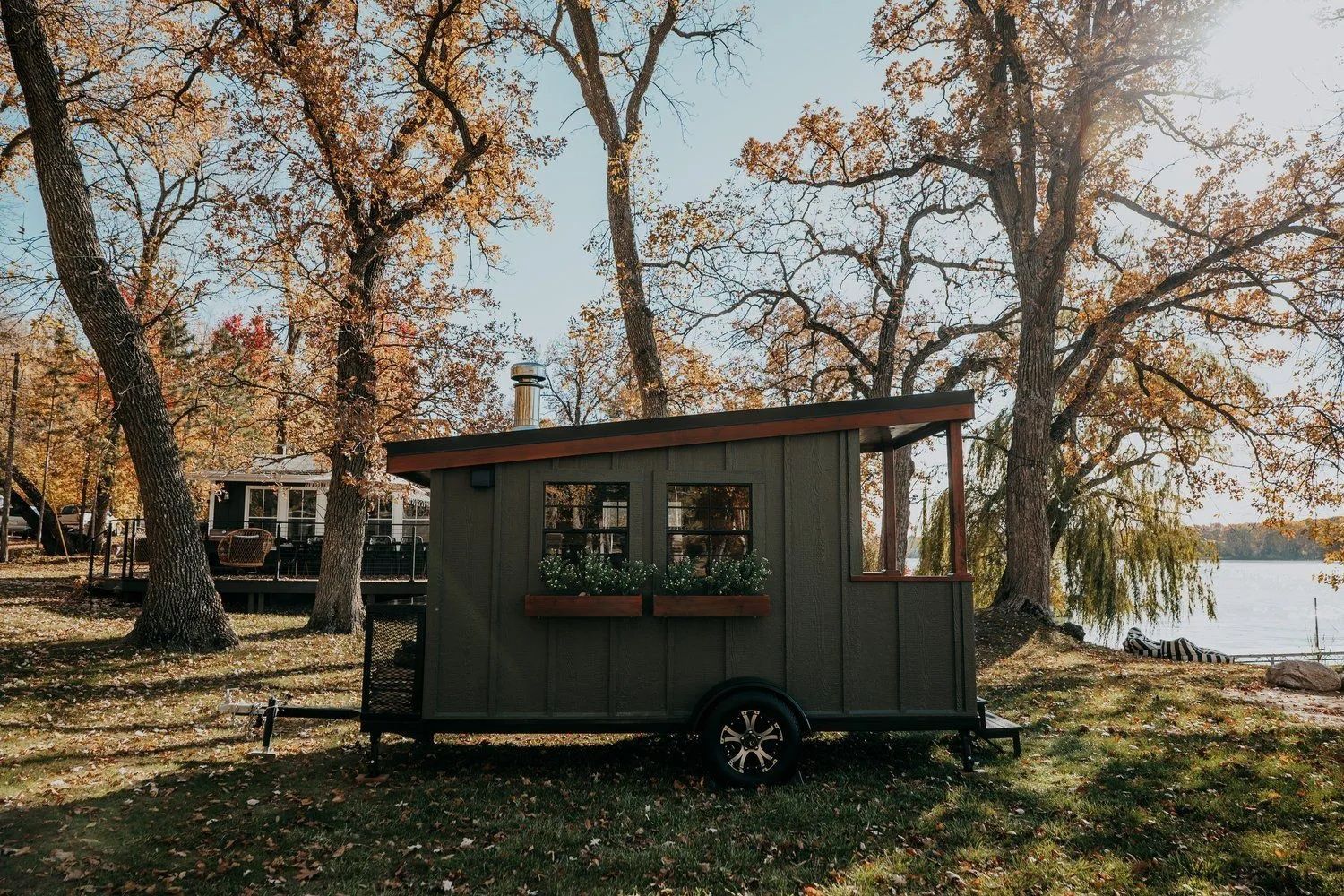 Tiny green cabin on a trailer with a window box, set in a grassy area near trees and a lake.