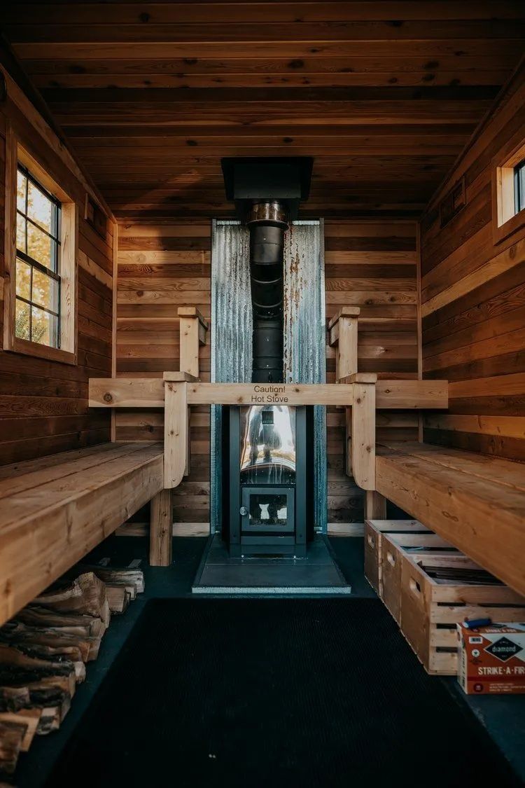 Interior of a small wooden cabin with a central fireplace, two benches, and small windows.